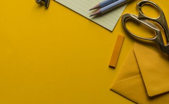 Gunting, amplop, dan pensil di atas latar kuning, ilustrasi KBLI ATK alat tulis kantor